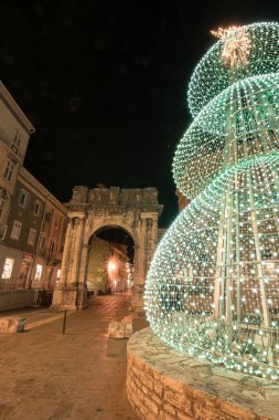 Kadim bir taş kemer ve yakınlardaki yapılar gece şenlikli süslemelerle parlayarak büyülü bir tatil atmosferi ve çarpıcı mimari tezatlıklar yaratıyor. Altın kapı, Pula..