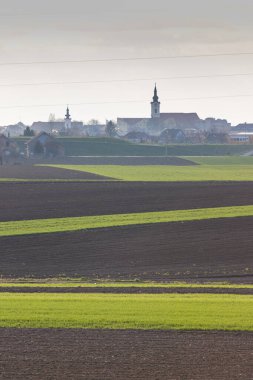 Uzakta büyüleyici bir köy ve kilise çan kuleleri olan, kırsal huzur ve doğal güzelliği yansıtan, açık tarım arazisi manzarası..
