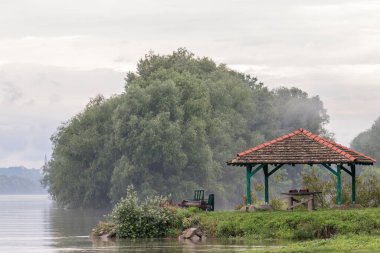 Güzel nehir manzaralı ahşap bir çardak, yemyeşil ağaçlar ve bulutlu gökyüzü..