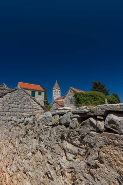 Idyllic rural scene showcasing a charming stone-built village featuring a church tower surrounded by greenery under a vivid blue sky. clipart