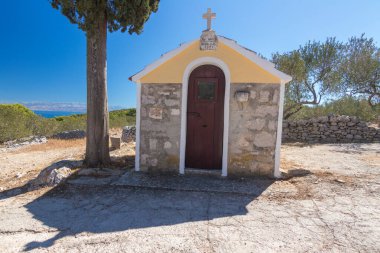 Uzun bir selvi ağacının yanında küçük bir şapel bulunan sakin Akdeniz manzarası canlı mavi bir deniz ve yemyeşil bir yer. Kompozisyon, Akdeniz mimarisi ve huzurlu doğal güzelliği yansıtıyor..