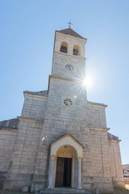 Ayırt edici bir çan kulesinin bulunduğu eski taş kilise, yemyeşil bir çan kulesiyle çevrili ve berrak, mavi bir gökyüzüne karşı güneş ışığıyla yıkanmış, dingin ve ruhani bir atmosferi resmediyor..