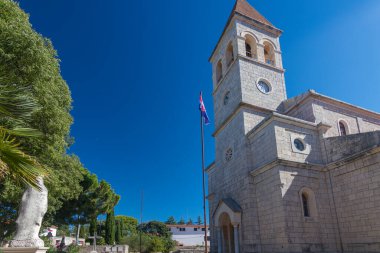 Ayırt edici bir çan kulesinin bulunduğu eski taş kilise, yemyeşil bir çan kulesiyle çevrili ve berrak, mavi bir gökyüzüne karşı güneş ışığıyla yıkanmış, dingin ve ruhani bir atmosferi resmediyor..
