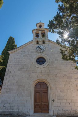 Ayırt edici bir çan kulesinin bulunduğu eski taş kilise, yemyeşil bir çan kulesiyle çevrili ve berrak, mavi bir gökyüzüne karşı güneş ışığıyla yıkanmış, dingin ve ruhani bir atmosferi resmediyor..
