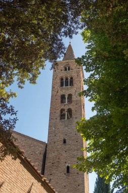 Kırmızı tuğlalarla inşa edilmiş antik bir kilise kulesi. Kemerli pencereler ve mimari detaylarla dolu. Parlak ve berrak bir gökyüzünün altında dimdik duruyor..