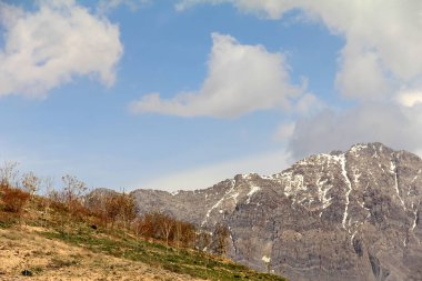 Görkemli kar örtülü dağlar parçalı bulutlu mavi bir gökyüzünün altında kurumuş çimenler tepenin önünü kaplıyor, doğanın ihtişamını ve huzur dolu güzelliğini sergiliyor..