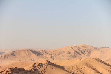 Geniş ve kurak bir çöl manzarası engebeli bir arazi ve parlak ve açık mavi bir gökyüzünün altında puslu bir dağ ufku sergiliyor. Sahne yalnızlık ve doğanın sonsuzluğu hissi yayıyor..