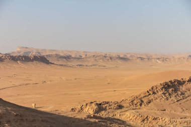 Geniş ve kurak bir çöl manzarası engebeli bir arazi ve parlak ve açık mavi bir gökyüzünün altında puslu bir dağ ufku sergiliyor. Sahne yalnızlık ve doğanın sonsuzluğu hissi yayıyor..