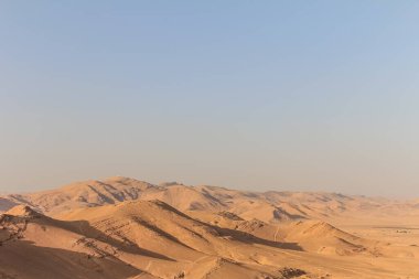 Geniş ve kurak bir çöl manzarası engebeli bir arazi ve parlak ve açık mavi bir gökyüzünün altında puslu bir dağ ufku sergiliyor. Sahne yalnızlık ve doğanın sonsuzluğu hissi yayıyor..