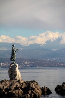 Rıhtımdaki bir kayanın üzerindeki bronz heykelin çarpıcı görüntüsü, geniş kıyı ve dağ manzarası, canlı bir gökyüzü altında, sanat ve doğal güzelliğin harmanlanmış hali..