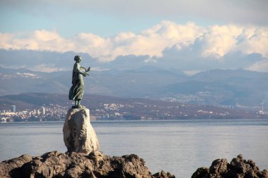 Rıhtımdaki bir kayanın üzerindeki bronz heykelin çarpıcı görüntüsü, geniş kıyı ve dağ manzarası, canlı bir gökyüzü altında, sanat ve doğal güzelliğin harmanlanmış hali..