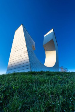Çembersel bir tasarıma sahip modern beyaz beton anıt, yeşil çevresi olan huzurlu bir semtte, berrak mavi bir gökyüzünün altında, kaldırım taşından yollar üzerine kurulmuş..