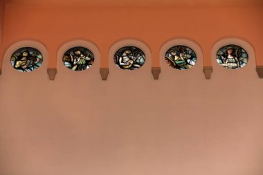 Zarif kemerli duvar, yumuşak sıcak ışıkla aydınlatılan, dört yuvarlak vitray pencereli, yumuşak dini temalı bir sanat eseri, kutsal bir kapalı alanda dingin ve ruhani bir atmosfer yaratıyor..