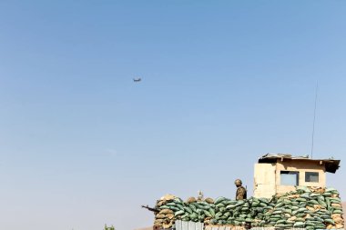 Mavi gökyüzünün altında nöbet tutan bir askerle etrafı kum torbalarıyla çevrili bir askeri gözetleme kulesi. Uzakta uçan bir helikopter görülüyor, sahnenin askeri ve savunma temasını vurguluyor.