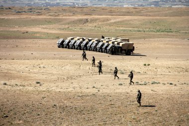 Üniformalı askerler geniş bir çöl ortamında konuşlanmış askeri malzeme kamyonlarının yanında yürürler..