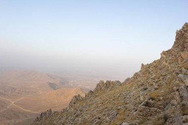 Yumuşak mavi bir gökyüzünün altında geniş bir manzaraya bakan kayalık bir yamaçtan büyüleyici bir manzara. Uzak arazi sisli bir ufuk oluşturuyor. Huzur ve macera hissi uyandırıyor..