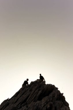 İki maceracının siluetleri sakin bir gökyüzünün altında engebeli bir kayalık tepeye tırmanıyor. Sahne takım çalışmasını, azmi ve gün batımının dingin parıltısında geçen açık hava keşiflerinin güzelliğini çağrıştırıyor..