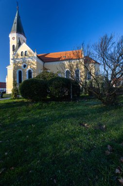 VELIKA GORİKA, CROATIA, 03.18.2020: Güneşli bir günde parkta uzun ağaçlarla çevrili huzurlu bir kilise. Sahne, doğal ortamdaki rahatlama veya yansıma için mükemmel sakin, resim gibi bir ortam yakalar..