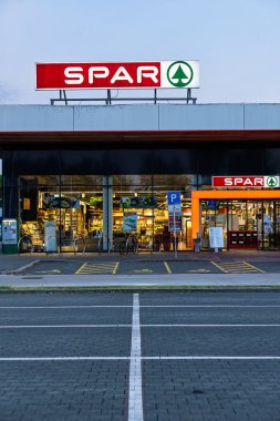 VELIKA GORICA, CROATIA, 04.22.2025: Aydınlatılmış bir market cephesinde müşteriler için elverişli kolaylıklar sunan geniş bir park alanı yer alıyor. Alacakaranlıkta yakalandı, karşılama ve organize perakende satışı sergiliyor..