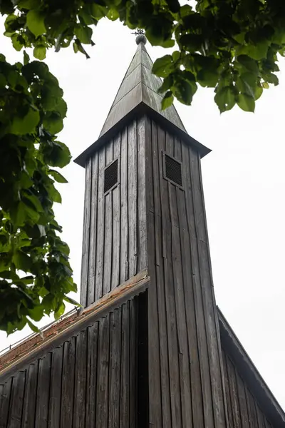Bereketli yeşil bir manzarada kırmızı kiremitli, ahşap bir kilise, sakin bir atmosferle çevrili. Bulutlu gökyüzü derinlik katıyor, doğaya karşı geleneksel mimari cazibeyi vurguluyor.