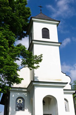 Tepesinde bir haç bulunan beyaz bir kilise kulesi mavi gökyüzünün altındaki yemyeşil ağaçlarla çevrili, duvarlarına yerleştirilmiş iki dini ikonu temsil ediyor, dindarlığı ve ruhani bağlılığı simgeliyor..