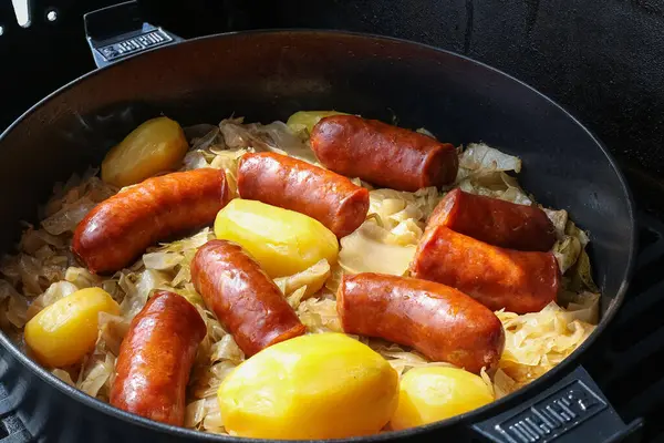 VINKOVCI, CROATIA, 04.07.2025: Lezzetli sosisler, bütün patatesler ve pişirilmiş lahana içeren ve tatmin edici bir yemek için bir tavada birlikte hazırlanan gerçek ve sağlıklı yemek.