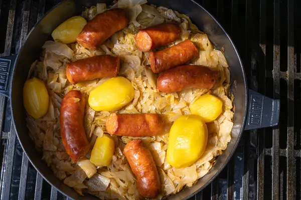 VINKOVCI, CROATIA, 04.07.2025: Lezzetli sosisler, bütün patatesler ve pişirilmiş lahana içeren ve tatmin edici bir yemek için bir tavada birlikte hazırlanan gerçek ve sağlıklı yemek.