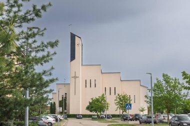 VELIKA GORICA, CROATIA, 04.23.2025: Çağdaş bir kilisenin dış görünüşü, üzerinde haç bulunan uzun bir saat kulesi. Park alanları ve ağaçlarla çevrili bulutlu gökyüzü huzurlu ve huzurlu bir şehir ortamı yaratıyor..