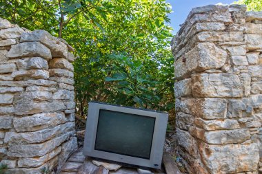 SOLTA, CROATIA, 08.21.2020: Parlak yeşil yeşillikler altında ve kırsal taş duvarlar arasında çürüyen ve doğal karşıtlık yaratan eski bir CRT televizyonu bırakıldı.