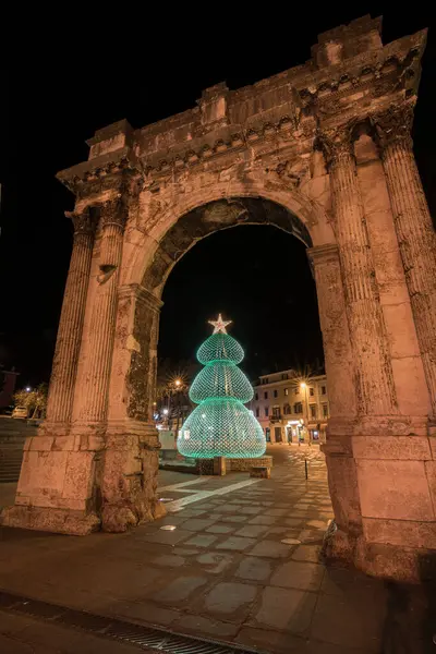 PULA, CROATIA, 01.06.2019: Kadim bir taş kemer ve yakınlardaki yapılar gece şenlikli dekorasyonlarla aydınlanarak büyülü bir tatil atmosferi ve çarpıcı mimari karşıtlıklar yaratıyor. Altın kapı, Pula..