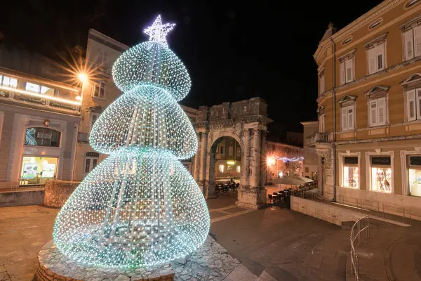 PULA, CROATIA, 01.06.2019: Kadim bir taş kemer ve yakınlardaki yapılar gece şenlikli dekorasyonlarla aydınlanarak büyülü bir tatil atmosferi ve çarpıcı mimari karşıtlıklar yaratıyor. Altın kapı, Pula..