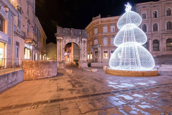 PULA, CROATIA, 01.06.2019: Kadim bir taş kemer ve yakınlardaki yapılar gece şenlikli dekorasyonlarla aydınlanarak büyülü bir tatil atmosferi ve çarpıcı mimari karşıtlıklar yaratıyor. Altın kapı, Pula..
