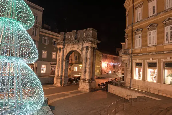 PULA, CROATIA, 01.06.2019: Kadim bir taş kemer ve yakınlardaki yapılar gece şenlikli dekorasyonlarla aydınlanarak büyülü bir tatil atmosferi ve çarpıcı mimari karşıtlıklar yaratıyor. Altın kapı, Pula..