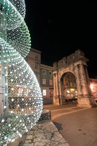 PULA, CROATIA, 01.06.2019: Kadim bir taş kemer ve yakınlardaki yapılar gece şenlikli dekorasyonlarla aydınlanarak büyülü bir tatil atmosferi ve çarpıcı mimari karşıtlıklar yaratıyor. Altın kapı, Pula..