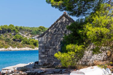 SOLTA, CROATIA, 08.28.2018: Sahil şeridindeki büyüleyici taş ev, canlı yeşil ağaçlar ve sakin bir turkuaz deniz ile çevrili, dinginlik ve doğal güzelliği aktarıyor..