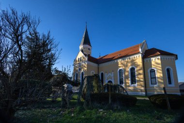 VELIKA GORİKA, CROATIA, 03.18.2025: Güneşli bir günde parkta uzun ağaçlarla çevrili huzurlu bir kilise. Sahne, doğal ortamdaki rahatlama veya yansıma için mükemmel sakin, resim gibi bir ortam yakalar..