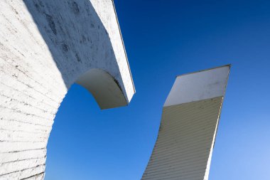 NOVA GORICA, CROATIA, 03.18.2025: Çembersel bir tasarıma sahip modern beyaz beton anıt, kaldırım taşlarının arasında, berrak mavi bir gökyüzünün altında yeşil çevreye sahip huzurlu bir semtte sergileniyor..