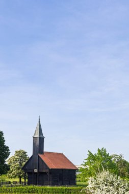 Yeşil tarlalar ve çiçek açan doğa, dinginlik ve miras cazibesi ile çevrili, sakin bir gökyüzünün altında kırmızı kiremitli bir çatı içeren tuhaf tarihi ahşap şapel..