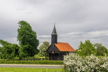 Yeşil tarlalar ve çiçek açan doğa, dinginlik ve miras cazibesi ile çevrili, sakin bir gökyüzünün altında kırmızı kiremitli bir çatı içeren tuhaf tarihi ahşap şapel..