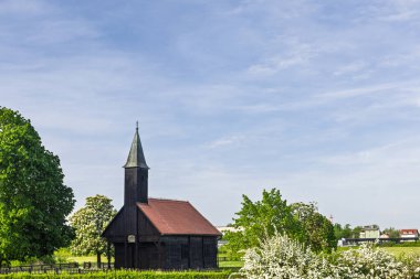 Yeşil tarlalar ve çiçek açan doğa, dinginlik ve miras cazibesi ile çevrili, sakin bir gökyüzünün altında kırmızı kiremitli bir çatı içeren tuhaf tarihi ahşap şapel..