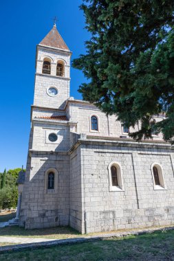 Tarihi bir taş kilisede uzun bir saat kulesi, kemerli pencereler ve fayanslı bir çatı bulunur. Mimari, gelenek ve barışçıl çevrenin altını çizen yemyeşil bir ağacı olan açık mavi gökyüzüne karşı..