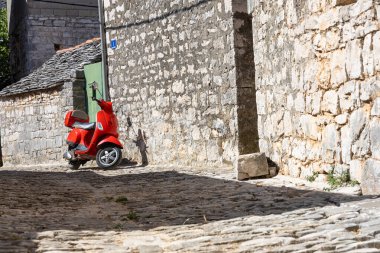Parlak kırmızı bir scooter, tarihi bir köyde taştan duvarlarla çevrili, güneşli bir cadde boyunca dinleniyor. Kırsal dokular, dar şeritler ve sıcak ışık resimli bir seyahat sahnesini çağrıştırıyor..