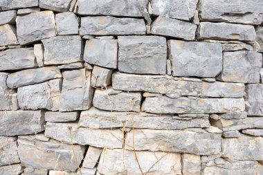 A weathered gray stone wall built from irregular rocks, showing layered masonry and hints of dry grass in the foreground. Ideal for rustic architecture, durability, and natural construction themes.