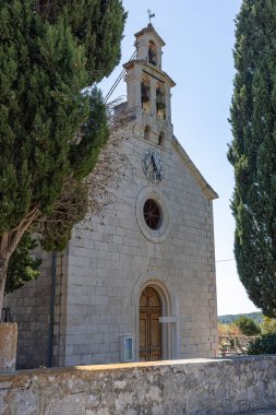 A small stone church featuring a clock face and twin bell towers, framed by two towering cypress trees against a bright blue sky. A serene, historic religious site in a rural setting.
