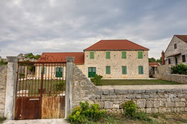 A stone two-story house with green wooden shutters sits in a quiet rural yard. Red-tiled roof, gravel path, and small shrubs create a calm, picturesque village scene.