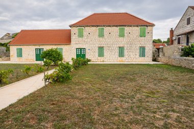 A stone two-story house with green wooden shutters sits in a quiet rural yard. Red-tiled roof, gravel path, and small shrubs create a calm, picturesque village scene.