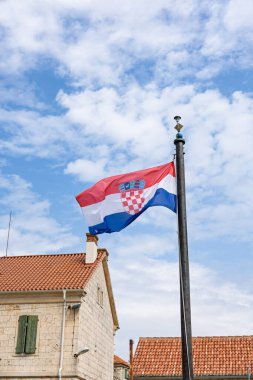 Taştan bir köyün üzerinde yeşil panjurlu ve kırmızı kiremitli çatılı parlak mavi gökyüzüne dikilmiş bir Hırvat bayrağı görülüyor. Kültürü ve mirası yansıtan sakin, kırsal bir manzara..