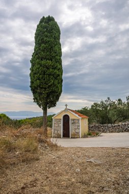 Sarı bukleli ve kırmızı kiremitli küçük bir taş şapel, güneşte pişirilmiş kırsal bir alanda, selvi çiçeğinin yanında yer alır. Sahne, dinginliği, yalnızlığı ve kırsal cazibeyi yansıtıyor..