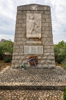 DONJE SELO, CROATIA, 08.24.2025: Bir heybetli taş savaş anıtı, ölen kahramanların anısına oyulmuş bir işçi figürü ve bir yıldız amblemi içerir. Üste bir çelenk, kaldırımlı bir meydanın ve bulutlu bir gökyüzünün altında alçak bir yeşilliğin ortasında duruyor..