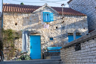 Canlı mavi kapıları ve panjurları olan yıpranmış taştan bir ev güneşli bir Akdeniz caddesinde oturuyor. Ivy duvara tırmanıyor, mavi bir bank cazibe katıyor, ve sahne kıyı köyünün sıcaklığını çağrıştırıyor..